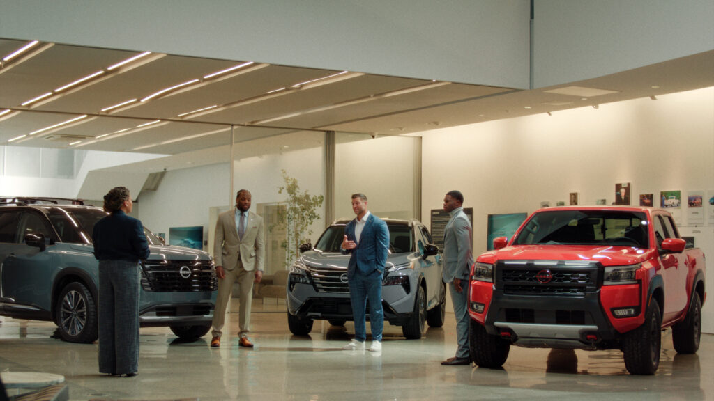 A group of people in business clothes, standing around three Nissan vehicles in a showroom.