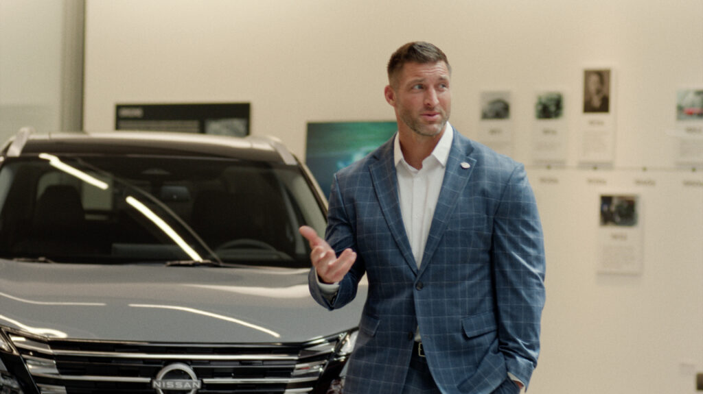 Tim Tebow in a plaid suit, standing in front of a Nissan Rogue, holding his hand up and looking to the right.